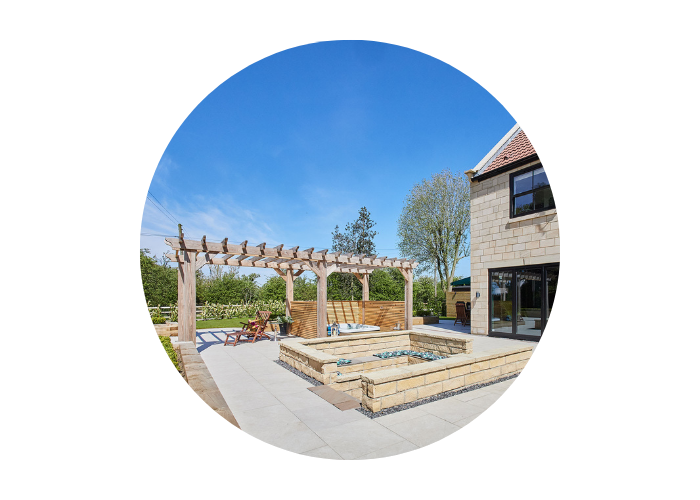 A view of the outdoor space in Finch House in Stokesley. The image shows a patio with white tiles, a light wood pergola above a hot tub, and a sunken seating space. 