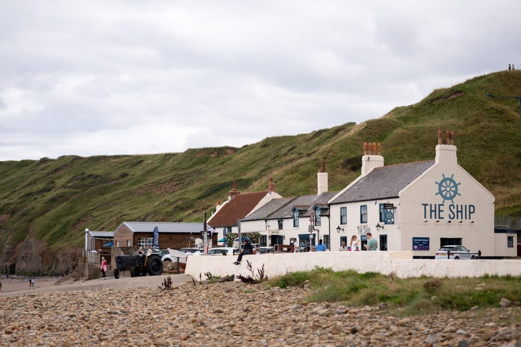 A view of The Ship Inn, Saltburn-by-the-Sea. The image shows a white building with grassy hills behind and a beach in front.