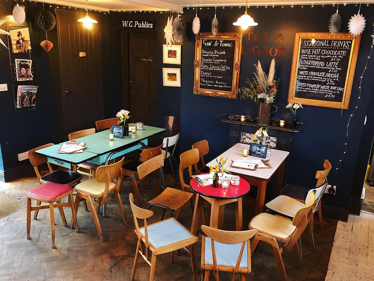 Tables at The Sitting Room, Saltburn-by-the-Sea. The walls are black with fairy lights and paper ornaments dangling from the ceiling. The tables are rustic with mis-matched wooden chairs around them. 