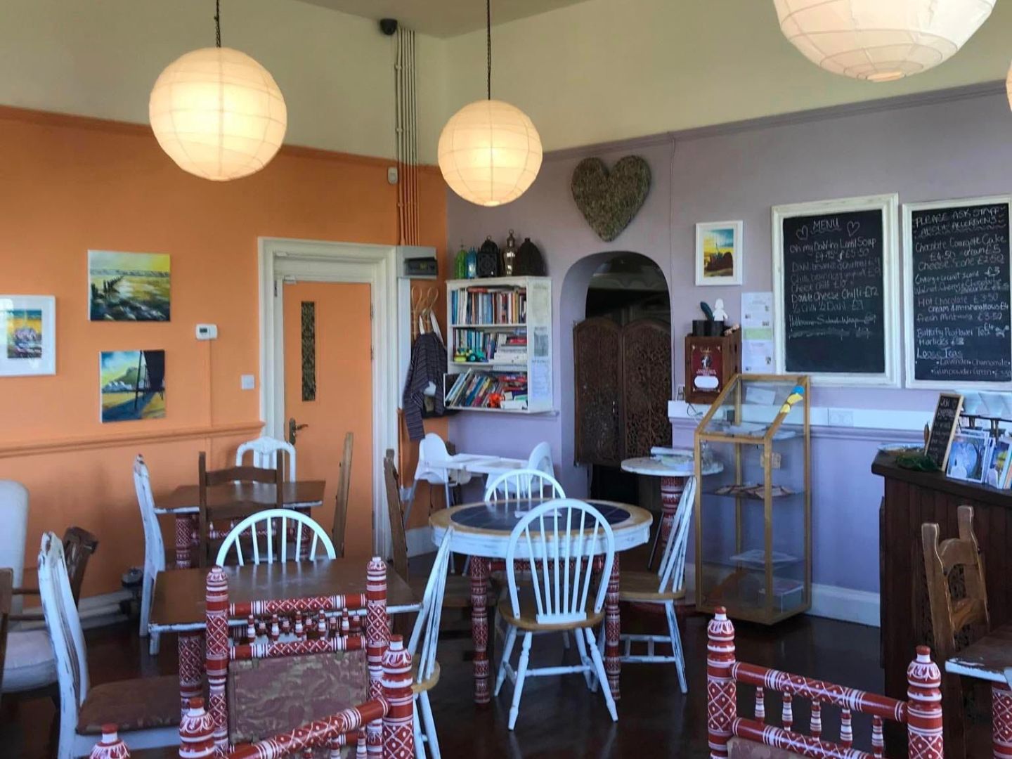 Tables at Shanti Cafe, Saltburn-by-the-Sea. The image shows a room with orange and lilac walls, and paper lanterns hanging from the ceiling. In the forefront of the image are mis-matched wooden tables and chairs. 