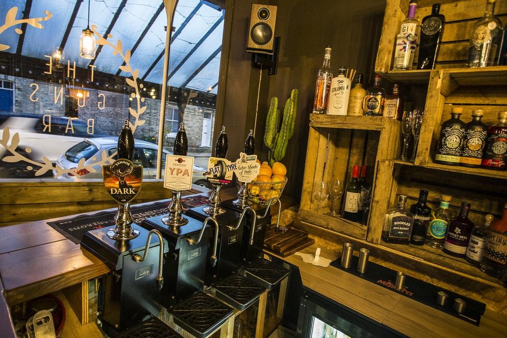 Inside The Guns Bar, Saltburn-by-the-Sea. The image shows a window in the background with The Guns Bar logo on in white. In the forefront of the image is the rustic wooden bar with draught pumps and bottles of spirits.