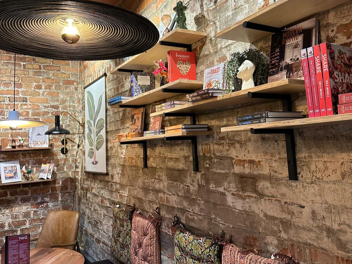 The decor in yard, Saltburn-by-the-Sea. The image shows exposed brick walls with shelves stacked with books, ornaments and plants. There are patterned cushions on the wall for seating. At the forefront is a large black light shade. 