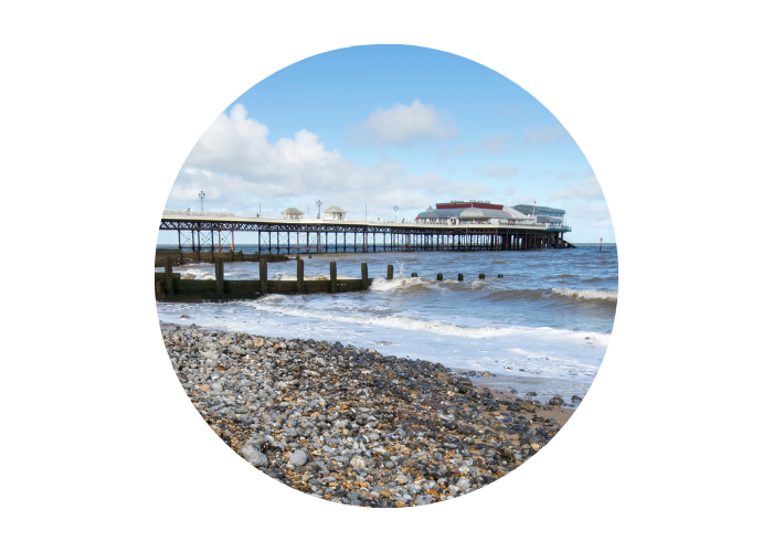 The image shows Cromer Pier from the pebble beach. The pier stretches out into the sea and there is a light blue sky above.