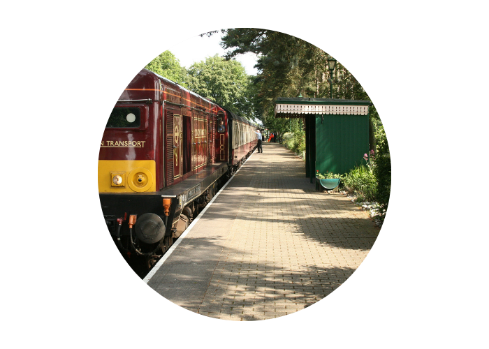 An image of Holt Train Station in Norfolk. There is a platform on the right with a dark green shelter. On the left-hand side of the platform is a traditional train in burgundy and yellow.